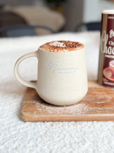 Load image into Gallery viewer, A personalized beige mug engraved with a cursive script on it, filled with a beverage, placed on a wooden coaster.