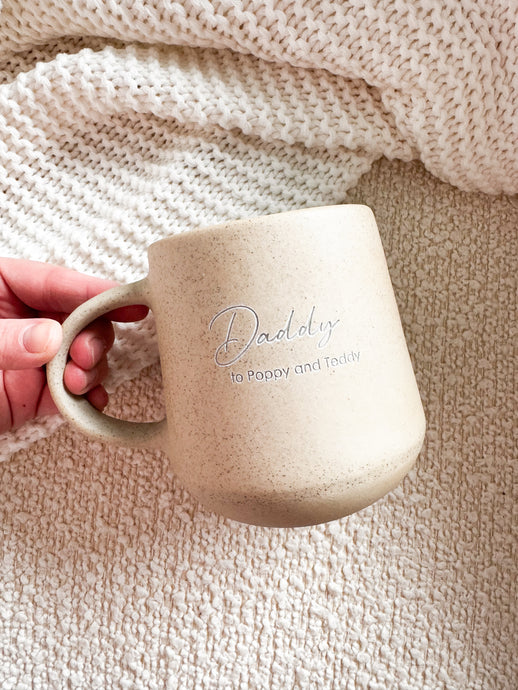 Beige ceramic mug with 'Daddy' engraving held by a hand against a textured beige background